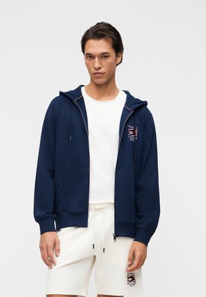 Giovane uomo che indossa una felpa con cappuccio blu navy con zip sopra una t-shirt bianca e pantaloncini bianchi con coulisse e loghi del marchio sul petto e sulla gamba.