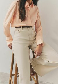 Chemise pêche rayée avec col à boutons, associée à un pantalon large beige taille haute. Ceinture marron simple et sandales neutres complètent le look.