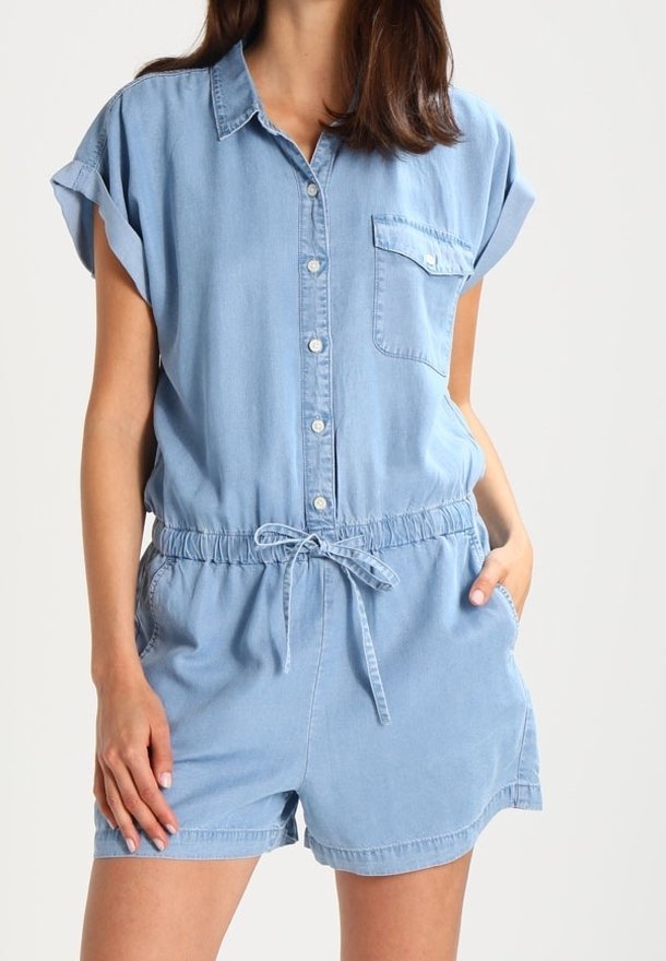 Femme portant une combinaison short en denim bleu clair à manches courtes, avec des boutons à l'avant, une poche poitrine, une taille à cordon de serrage et des poches latérales.