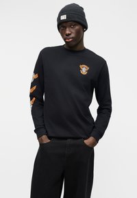 Maglietta nera a maniche lunghe con logo grafico arancione sul petto e sulla manica; abbinata a pantaloni neri e a un berretto lavorato a maglia grigio scuro.