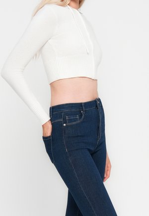 Femme portant un sweat à capuche court blanc et un jean taille haute bleu foncé, avec une main dans la poche, vue des épaules aux hanches.