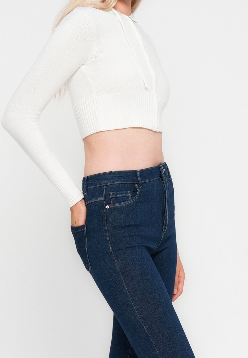 Femme portant un sweat à capuche court blanc et un jean taille haute bleu foncé, avec une main dans la poche, vue des épaules aux hanches.