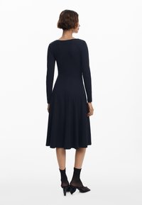 Schwarzes Kleid mit langen Ärmeln, körperbetontem Oberteil und ausgestelltem Rock. Glatter Stoff, Bootsausschnitt und knielanger Saum. Getragen mit durchsichtigen schwarzen Socken und hohen Schuhen.