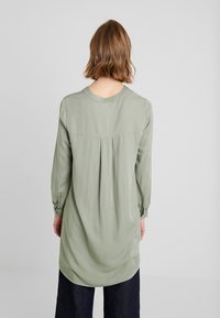 Langärmlige, hellgrüne Tunika mit rundem Halsausschnitt, Falten auf der Rückseite und elastischen Bündchen. Der Stoff wirkt glatt und fließend.