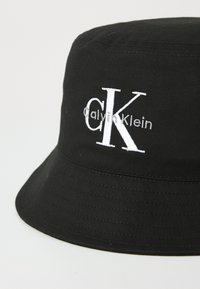 Black bucket hat made of fabric featuring a white embroidered Calvin Klein logo; structured shape with a wide, downward-sloping brim.