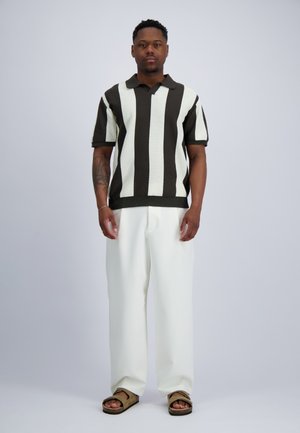 Man die staat en een zwart-wit verticaal gestreept shirt met korte mouwen, witte broek en bruine sandalen draagt op een grijze achtergrond.
