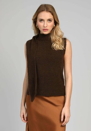 Femme portant un haut sans manches en tricot marron foncé avec un détail en écharpe et une jupe en satin couleur cuivre, debout devant un fond gris uni.