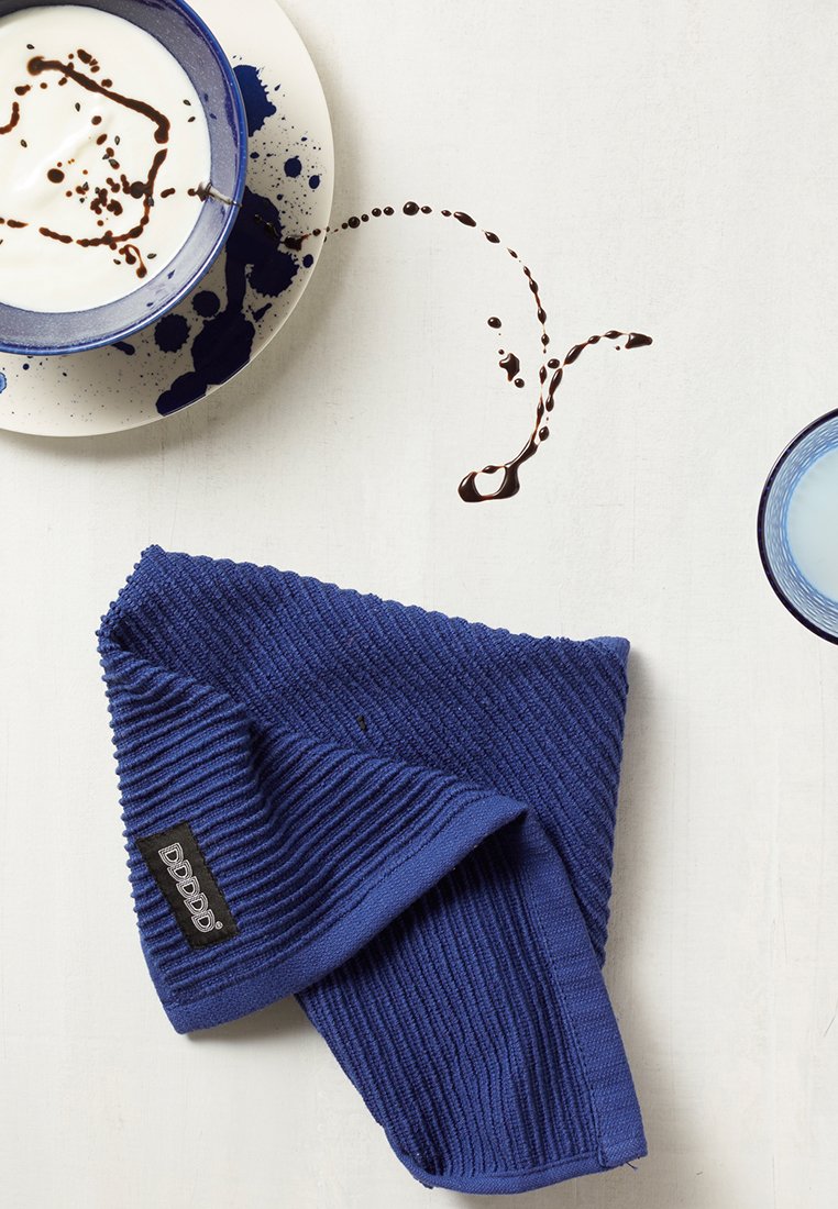 Blau strukturiertes Tuch neben einer blauen Tasse und Untertasse mit verschütteter dunkler Flüssigkeit auf einer weißen Oberfläche.