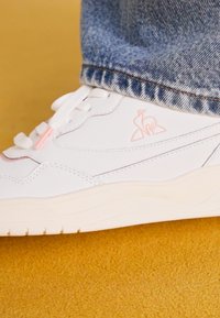 Fehér sneaker sima bőr felsőrésszel, diszkrét rózsaszín logóval és kontrasztos varrással. Hullámos gumi talp és kerek orr design. Farmer szegély látható.