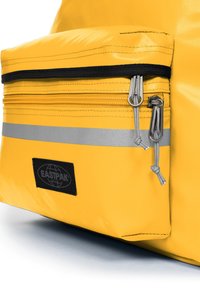 Sac Eastpak jaune avec une surface texturée, une poche zippée à l'avant avec des accessoires en argent et une étiquette logo noire. Détail de bande réfléchissante présent.