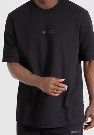 Mężczyzna noszący jednokolorowy czarny t-shirt z krótkim rękawem z małym logo "SMILODOX" na piersi i metką na dolnej stronie boku.