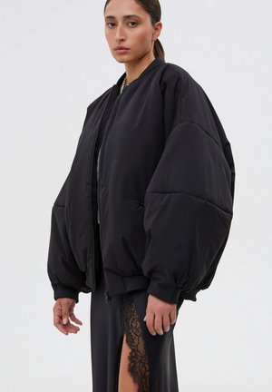 Kobieta w dużej czarnej puchowej kurtce na zamek oraz czarnej spódnicy z koronkowym rozcięciem do uda, stojąca na tle jasnego, jednolitego tła.