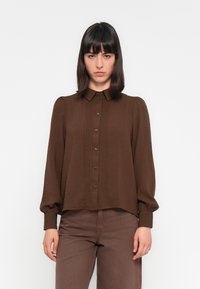 Camisa castanha de botão, com um tecido texturizado, apresentando frente plissada, mangas longas e bufantes, e um colarinho clássico.