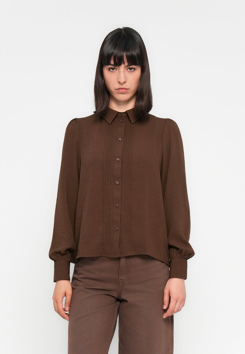 Camisa castanha de botão, com um tecido texturizado, apresentando frente plissada, mangas longas e bufantes, e um colarinho clássico.