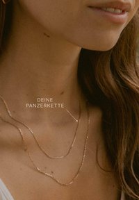 Gestapelte goldene Halsketten zeigen ein glattes, rundes Design mit unterschiedlichen Kettengliedern und Texturen, die auf einem nackten Hals ruhen.