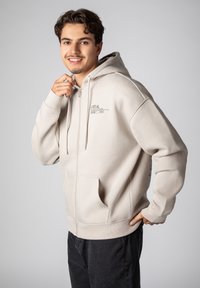Light beige zip-up hoodie in soft fabric, featuring a front pocket, and printed text on the chest. Hooded design with long sleeves.