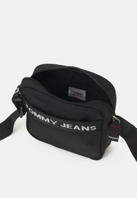 Bolso cruzado de tela negra con cierre de cremallera, que presenta el logo "TOMMY JEANS" en plateado y una correa ajustable. El interior no tiene forro.