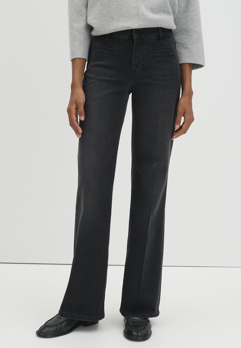 Jean en denim noir évasé avec une texture lisse, design à cinq poches et coutures contrastantes. Porté avec un pull gris clair et des chaussures noires.