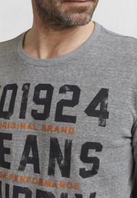 Grå T-shirt med stort sort trykt tekst og orange detaljer. Blødt stof med korte ærmer og en rund halsudskæring.