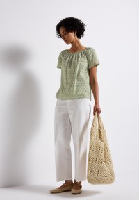 Blusa verde chiaro con scollatura arricciata e motivi a occhio, abbinata a pantaloni bianchi a gamba dritta e una borsa intrecciata beige.