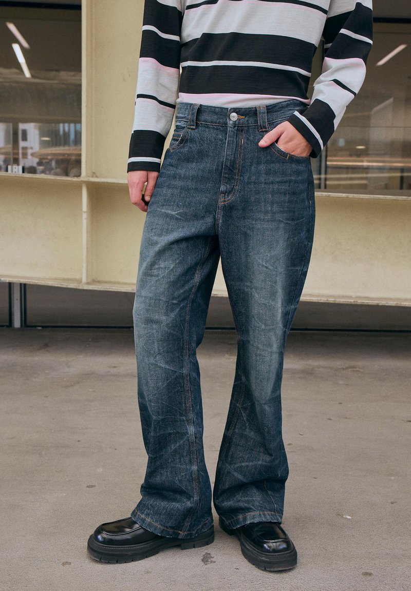 Jeans in denim blu scuro con una vestibilità rilassata, caratterizzati da cuciture arancioni, tasche laterali e uno stile delle gambe leggermente svasato.