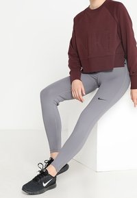 Sudadera corta color marrón oscuro con el logo texturizado de "NIKE", leggings ajustados grises con el swoosh negro de Nike, y zapatillas deportivas negras con cordones.
