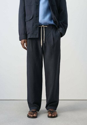 Hombre con pantalones negros holgados con cordón, una camisa azul, una chaqueta oscura y chanclas negras, de pie frente a un fondo liso.