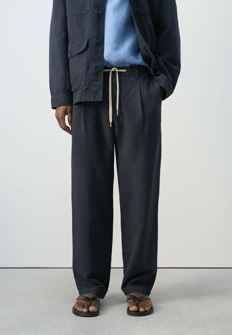 Homme portant un pantalon noir ample avec un cordon, une chemise bleue, une veste foncée et des tongs noires, debout devant un fond uni.