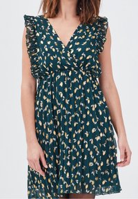 Robe verte avec un décolleté en V profond, des manches à volants, et un motif floral avec des accents dorés et blancs. Tissu léger et plissé.