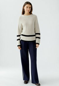 Cremfarbener Strickpullover mit schwarzen horizontalen Streifen und Knopfdetails an der Seite, kombiniert mit maßgeschneiderten marineblauen Hosenträgern und schwarzen Schuhen.
