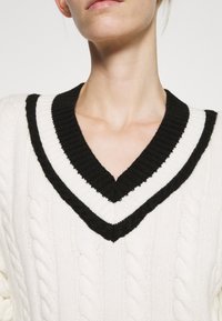 Pletený svetr se výstřihem do V, s černo-bílým žebrovaným límcem. Tělo svetru má krémový copánkový vzor.