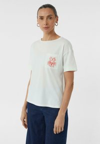 T-shirt blanc à manches courtes avec une poche ornée d'une broderie de homard rouge. Tissu lisse avec une coupe décontractée, associé à un jean large bleu marine.