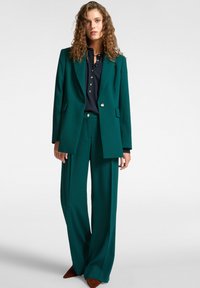 Completo verde acqua con giacca strutturata e pantaloni a gamba larga, abbinato a una camicia scura con bottoni. Presenta dettagli con bottoni dorati e una texture liscia.