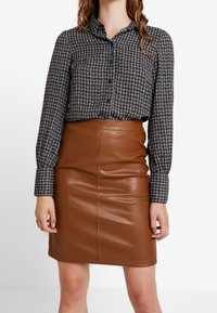 Blusa a quadri con maniche lunghe e chiusura a bottoni, abbinata a una gonna in pelle marrone aderente con cuciture verticali.