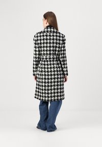 Vilnas maisījuma mētelis ar melnbaltu houndstooth rakstu, jostots viduklis un noklājuma apkakle. Garas piedurknes ar precīzu piegriezumu.