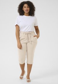 Pantalons beiges taille haute avec une ceinture en tissu, dotés de poches latérales et de revers. Associés à un t-shirt blanc uni et des sandales à talons blancs.