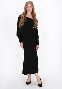 Bluză neagră, cu mâneci lungi, off-the-shoulder, combinată cu o fustă lungă, mulată, asortată. Materialul pare neted și moale. Pantofii stiletto negri completează look-ul.