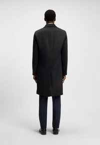 Manteau en laine noir avec des revers crantés et une coupe droite, affiché de dos, associé à un pantalon sombre et des chaussures noires.