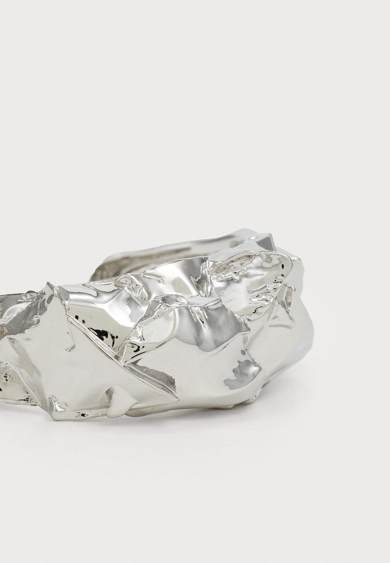 Gekreukeld zilveren metalen armband, met een gladde textuur en onregelmatige vorm, voorzien van glanzende oppervlakken en duidelijke kreukdetails.
