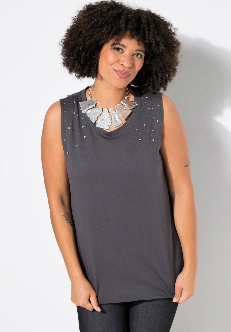 Top grigio senza maniche con accentuazioni borchiate sulle spalle, abbinato a una grande collana metallica. Tessuto morbido e drappeggiato; vestibilità rilassata.