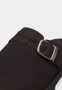 Κοντινή λήψη λουριού παπουτσιού από σκούρο καφέ σουέντ με ασημένιο μεταλλικό αγκράφα σε απλό φόντο.