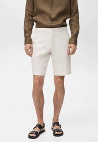 Pantaloni corti in lino color beige chiaro con taglio dritto, dotati di chiusura con un solo bottone. Indossati con una camicia a maniche lunghe marrone scuro e sandali neri.