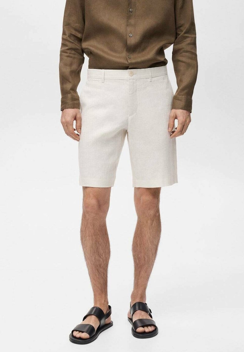 Pantaloni corti in lino color beige chiaro con taglio dritto, dotati di chiusura con un solo bottone. Indossati con una camicia a maniche lunghe marrone scuro e sandali neri.