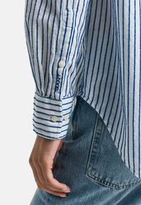 Wit overhemd met blauwe verticale strepen. Voorzien van een manchetknoop en een logo. Gekoord met lichtblauwe denim, waarbij de stikseldetails zichtbaar zijn.