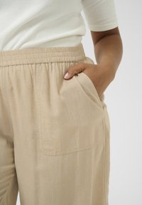 Pantalons beige clair, amples, avec une taille élastique et des poches latérales ; le tissu semble doux avec une texture subtile et un design léger.