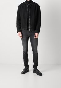 Chaqueta de mezclilla negra con cuello alzado, cierre frontal de cremallera y botón a presión. Combinada con jeans gris oscuro y mocasines negros.