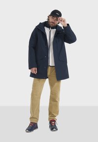 Giacca impermeabile blu navy con cappuccio, tasche frontali e chiusura a scatti, sovrapposta a una maglietta bianca, abbinata a pantaloni in corduroy beige e scarpe sportive.