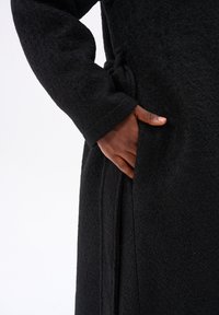 Czarny, teksturowany płaszcz z długimi rękawami i boczną kieszenią; posiada zawiązywany pasek do zapinania. Materiał wydaje się miękki i ciepły.