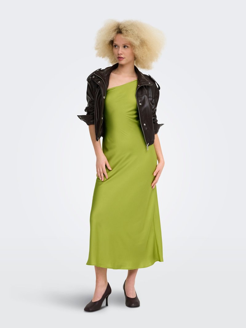 Femme aux cheveux blonds bouclés portant une robe verte à une épaule, une veste en cuir noire et des talons hauts noirs, debout devant un fond uni.