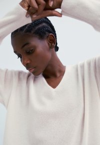 Junge Frau mit geflochtenem Haar, trägt einen hellbeigen V-Ausschnitt-Pullover, hebt ihre verscrönten Arme über ihren Kopf vor einem weißen Hintergrund.